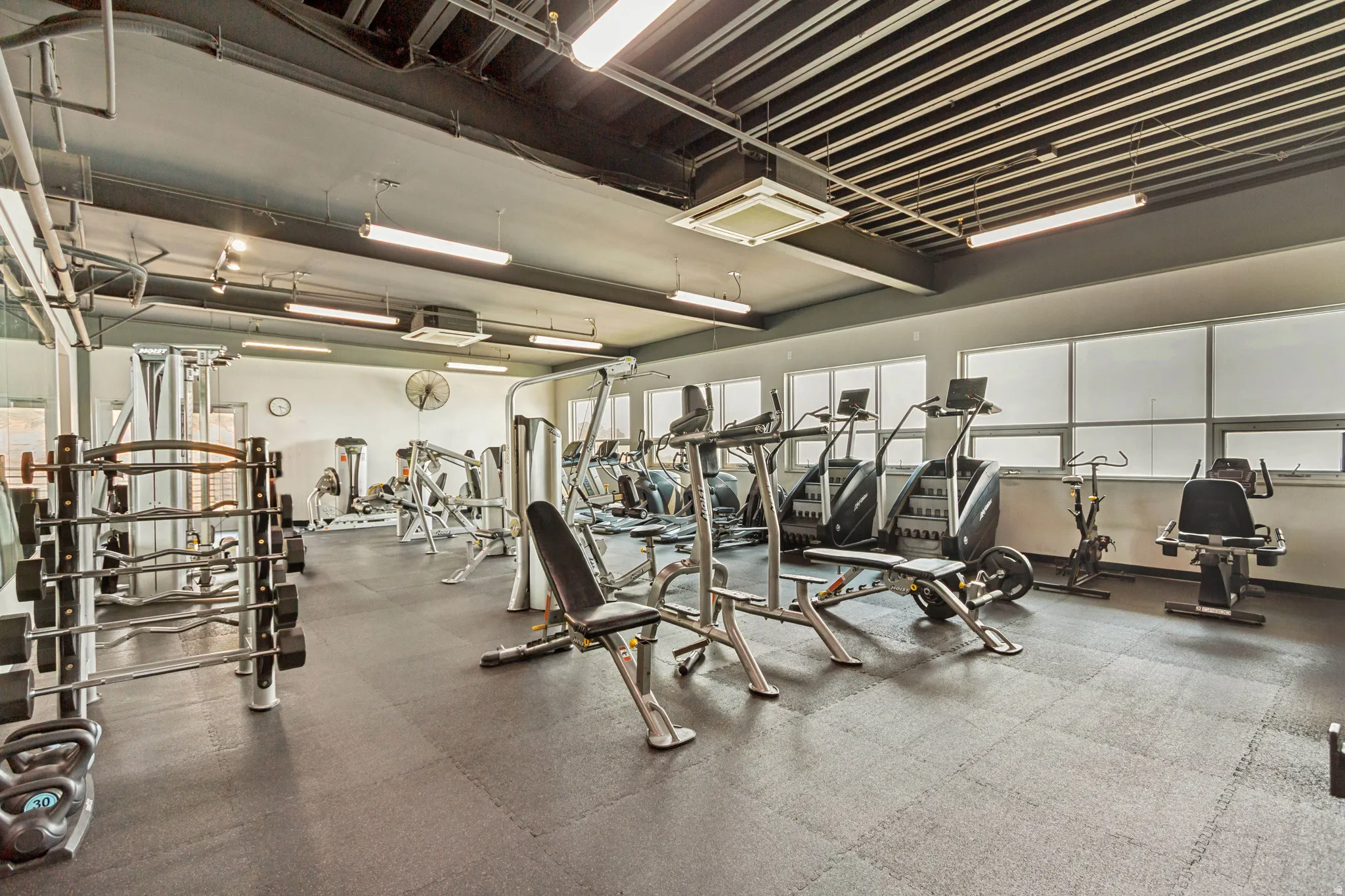 Fitness facility with extensive exercise equipment, rubber flooring, industrial ceiling, fluorescent lighting, and natural light from multiple windows