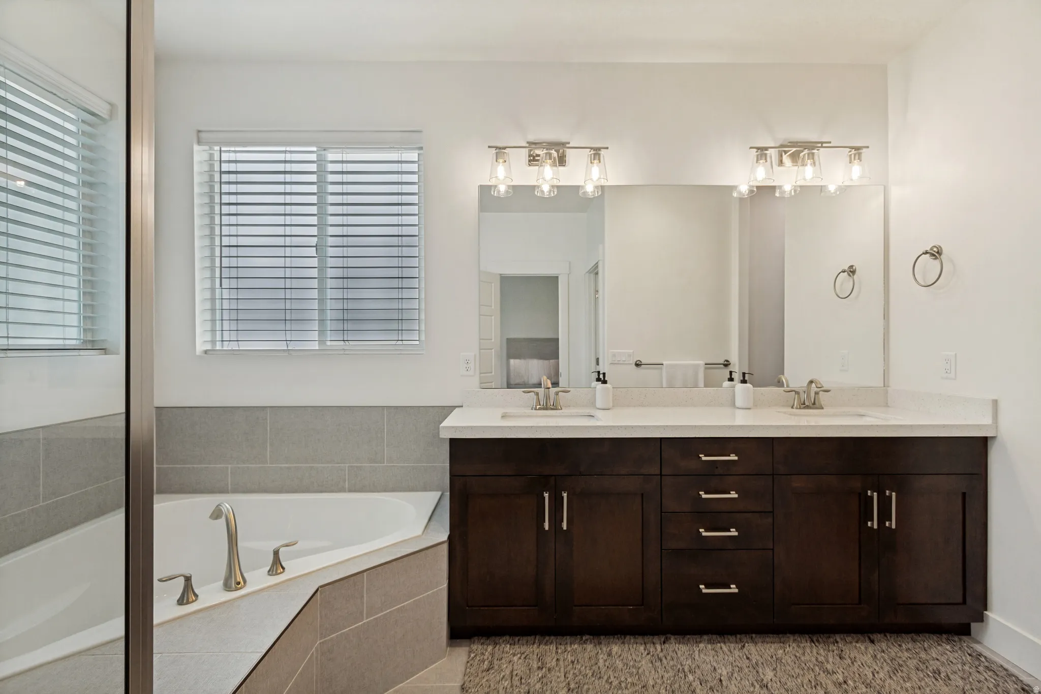 Spacious bathroom featuring a corner soaking tub with tile surround, a dual vanity with a light-toned countertop, brushed nickel fixtures, and decorative lighting