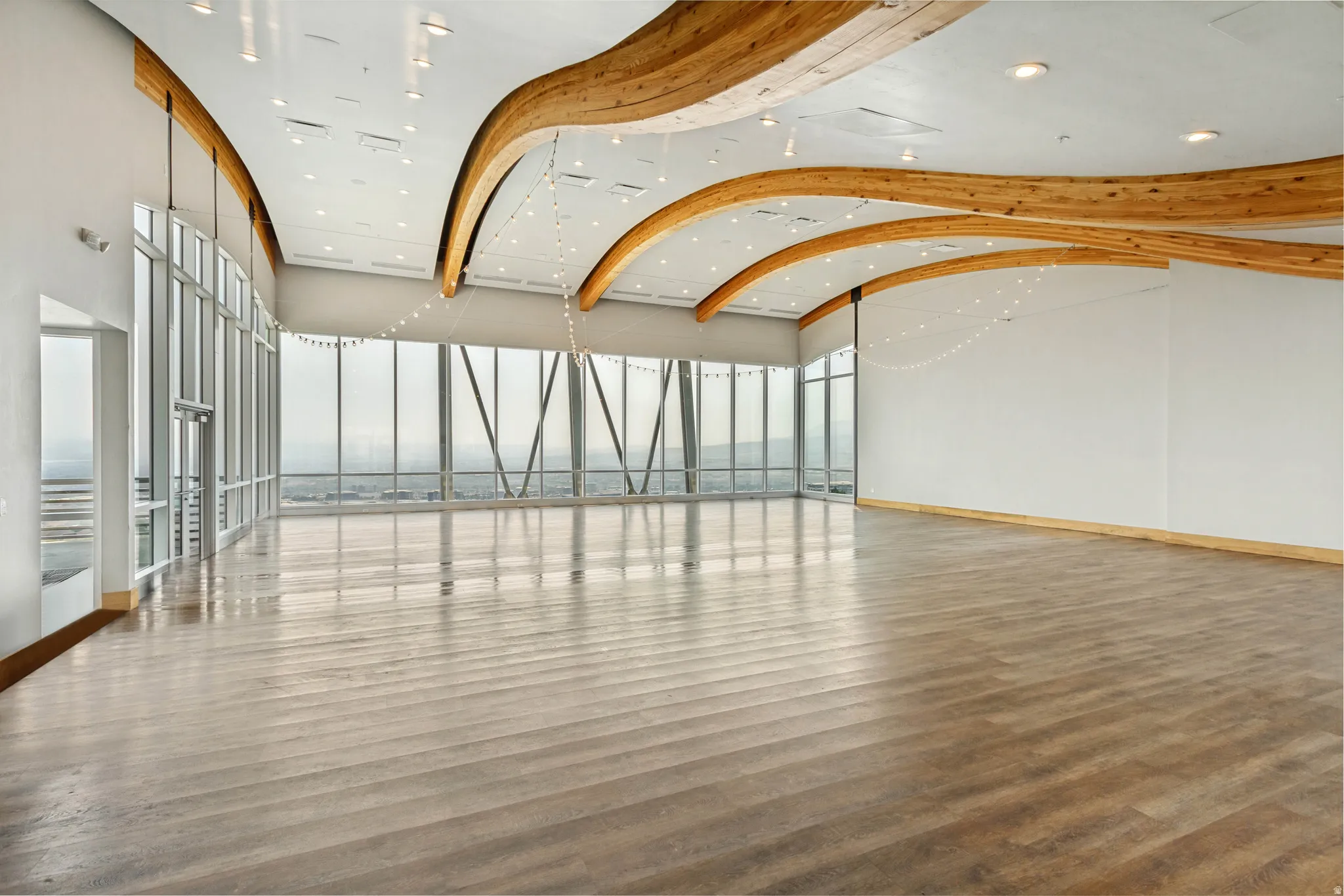 Spacious open-concept room featuring extensive window walls, exposed wooden ceiling beams, recessed lighting, and light wood-finish flooring