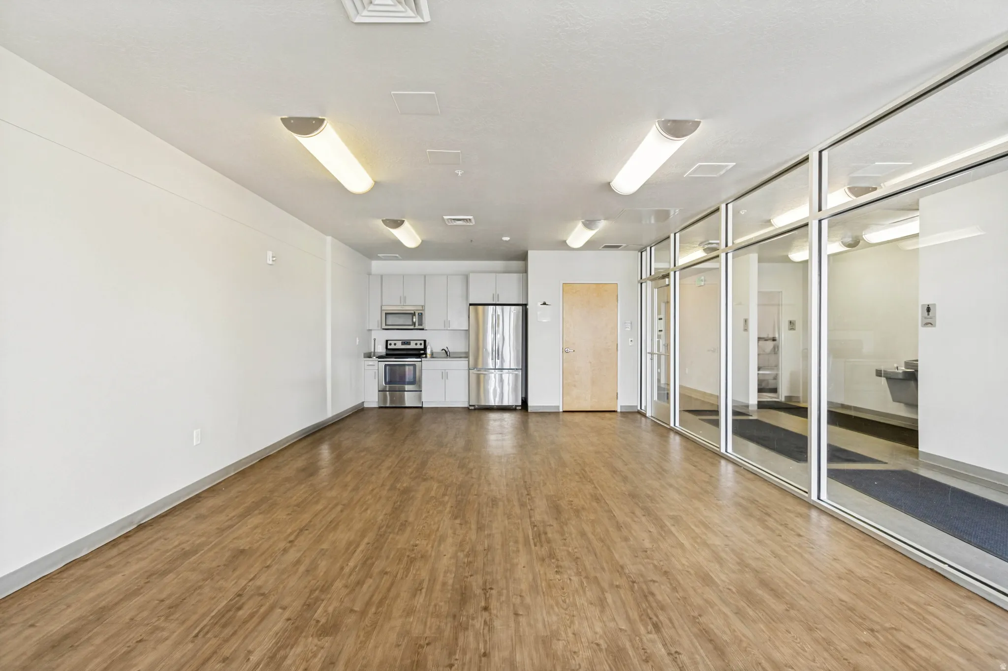 Spacious open-concept area featuring wood-finish flooring, a contemporary kitchenette with stainless steel appliances and light cabinetry, ample overhead lighting, and extensive full-height interior glass partitions