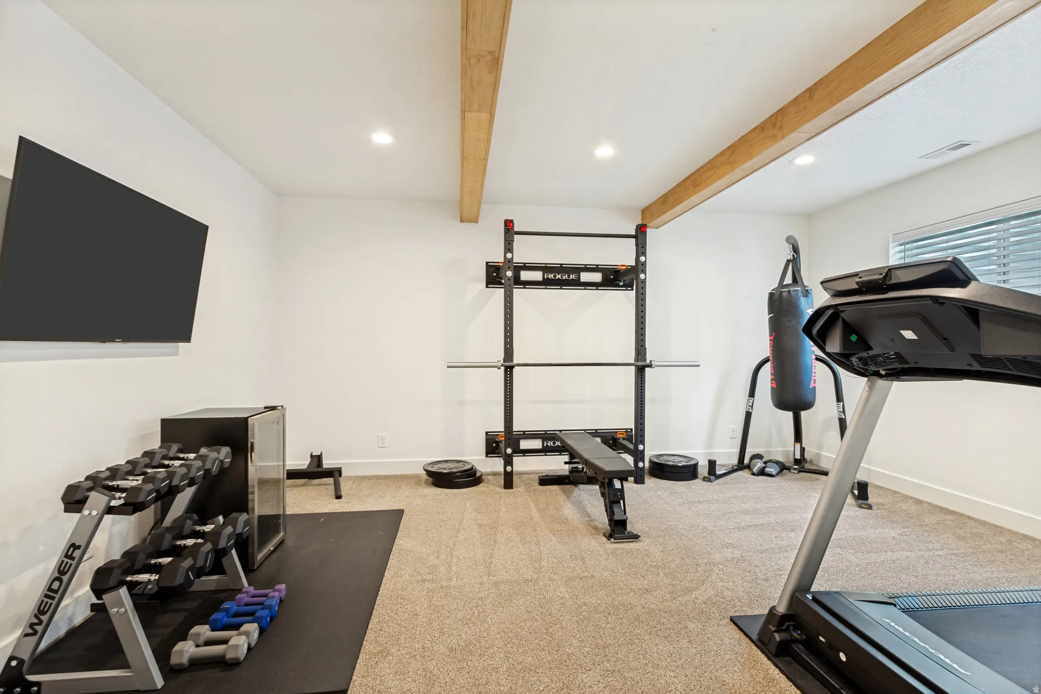 Dedicated home gym area featuring recessed lighting, exposed wood ceiling beams, wall-mounted television, and a window with blinds