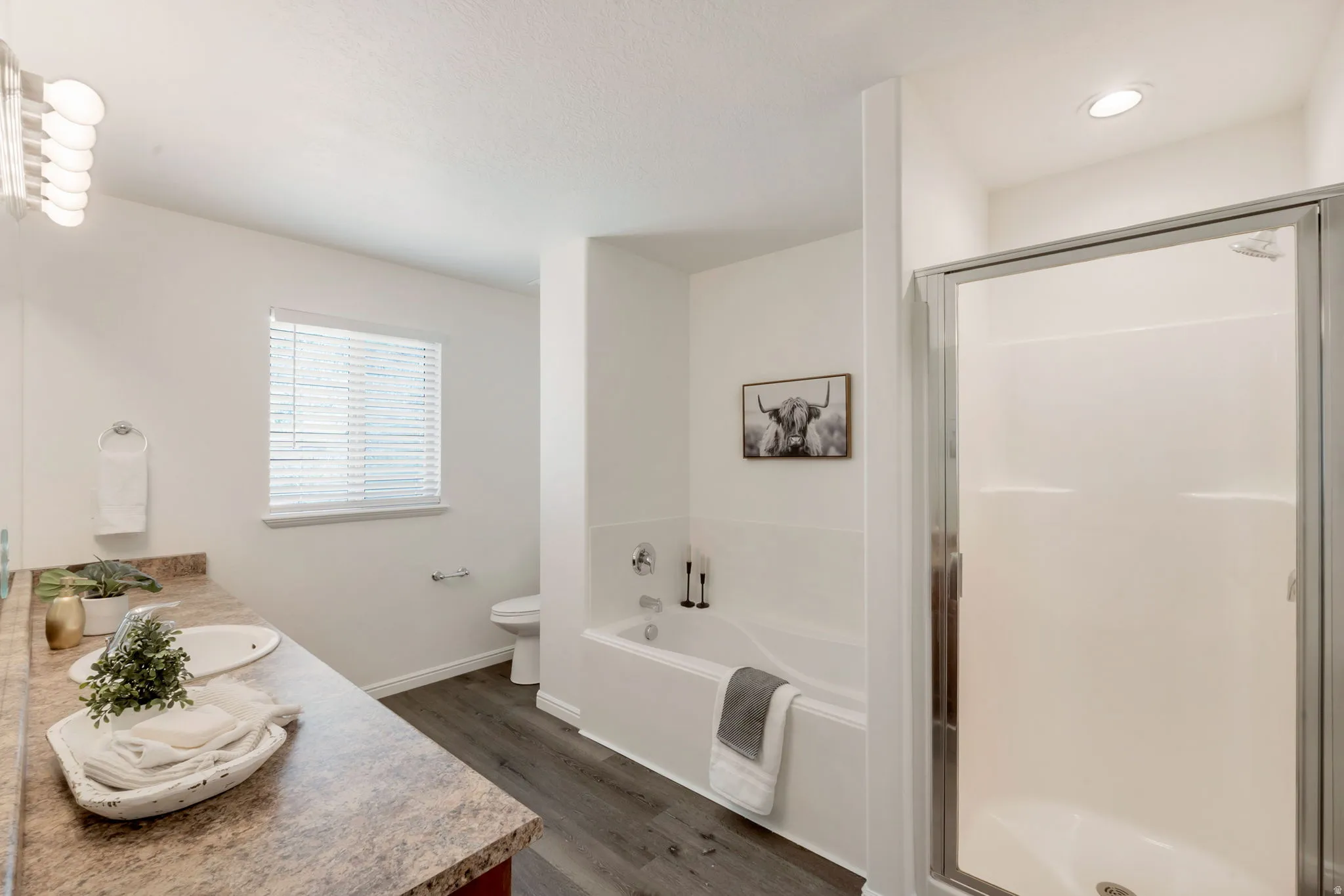 Bathroom featuring a dual vanity with a speckled countertop, a built-in soaking tub, a stand-up shower with a glass enclosure, and wood-finish flooring
