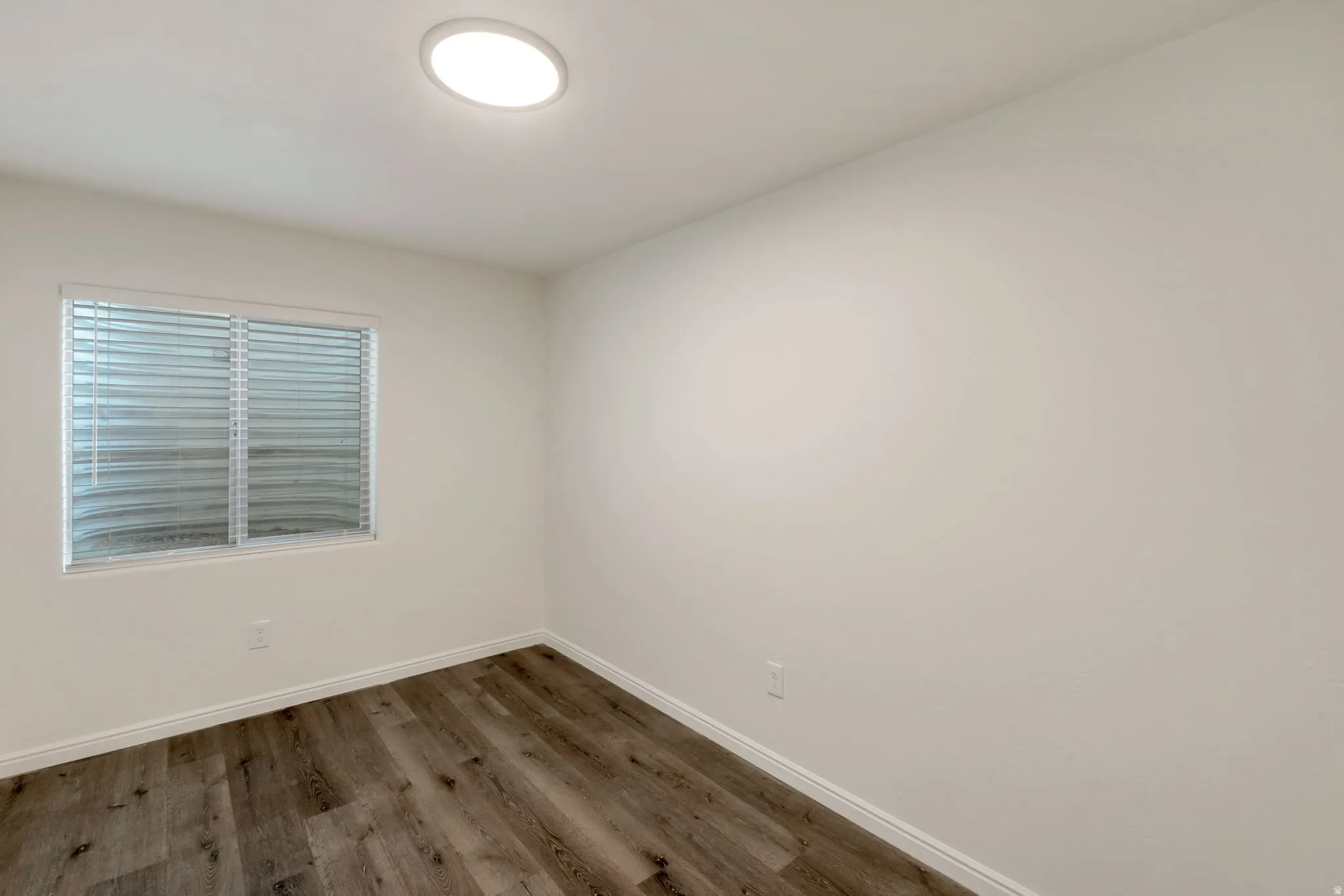 Room featuring wood-finish flooring, white baseboards, a single window with blinds, and a flush-mount ceiling light