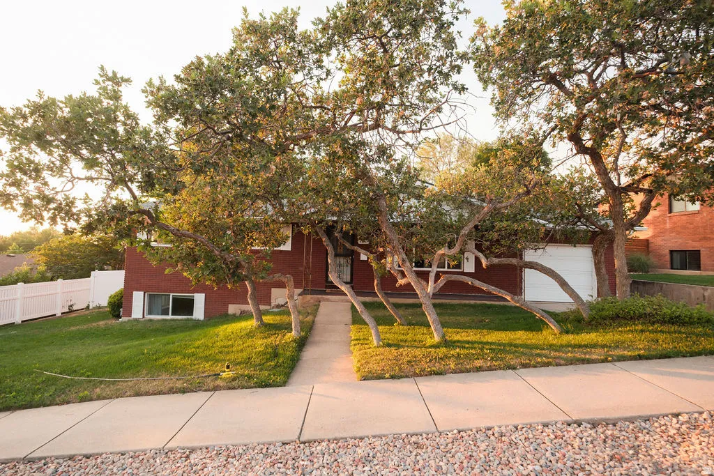 Mature trees frame the red brick exterior, featuring a white garage door, concrete walkway, and a white picket fence