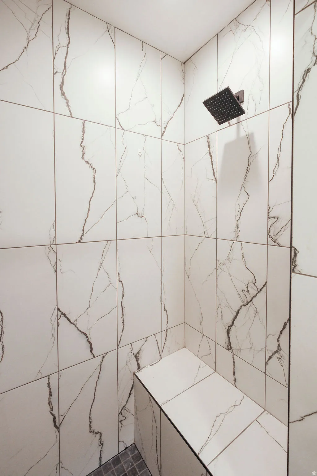 Shower stall featuring large format white tile with dark veining, a matte black rain shower head, a built-in bench, and dark square floor tiles