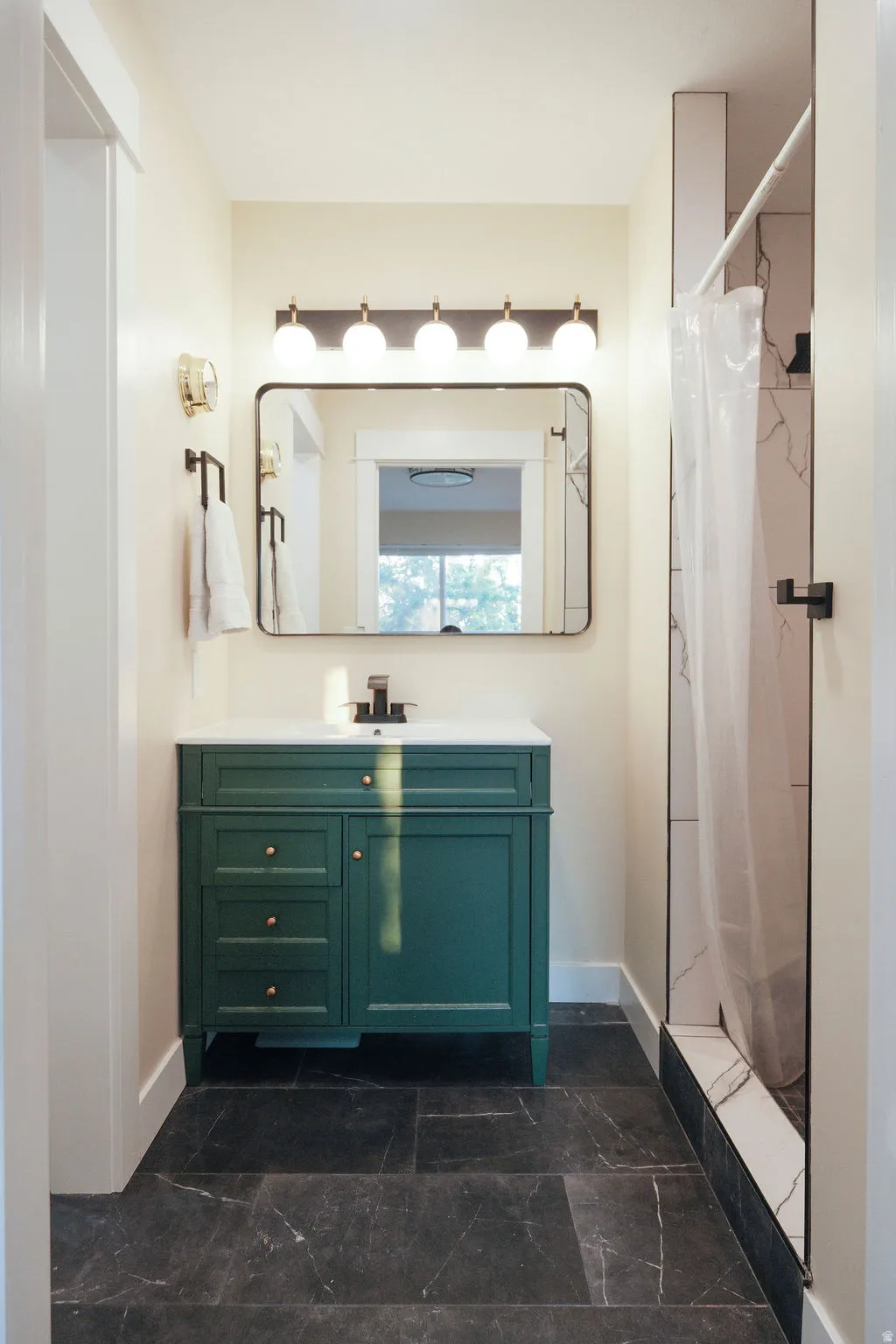 Dark gray tiled flooring with white veining, a deep green vanity with a white countertop and gold-toned hardware, a rectangular mirror, and a linear vanity light fixture with five spherical bulbs