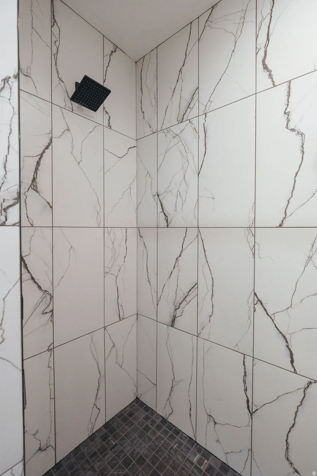 Shower stall featuring large format white tiled walls with dark veining, a matte black rain showerhead, and dark mosaic tile flooring