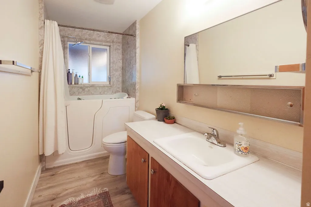 Bathroom featuring a walk-in tub with shower, wood-finish flooring, a vanity with integrated sink, and an expansive mirror
