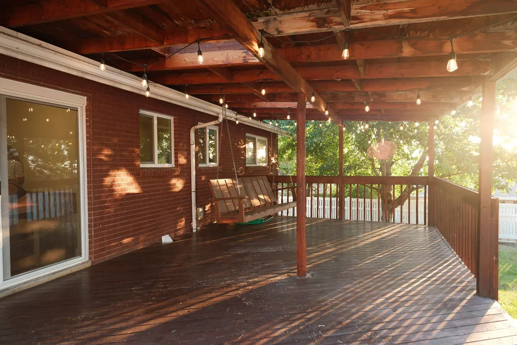 Expansive covered wood deck featuring a rich stain finish, exposed wood beam ceiling, decorative string lighting, and a white perimeter fence
