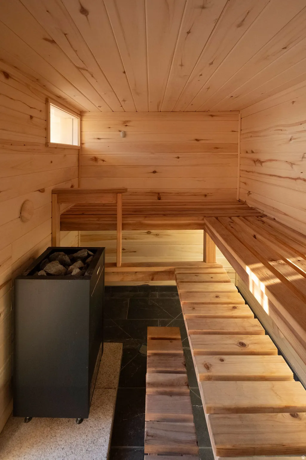 Built-in sauna featuring light wood paneling, multi-level wooden benches, a compact window, and a dark stone heater