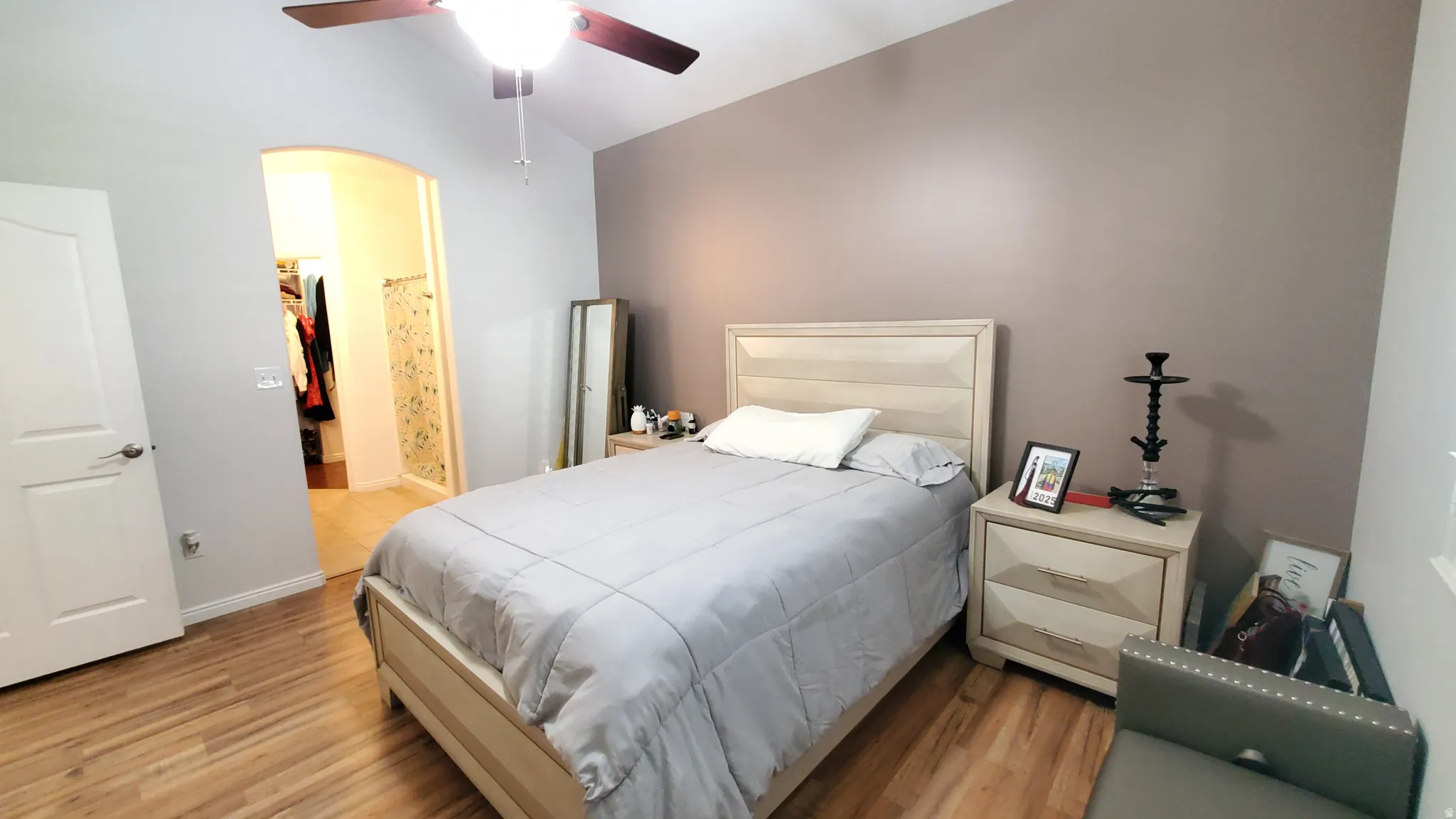 Vaulted ceiling bedroom featuring wood-finish flooring, a ceiling fan with integrated lighting, a neutral-toned accent wall, and an arched doorway leading to a walk-in closet