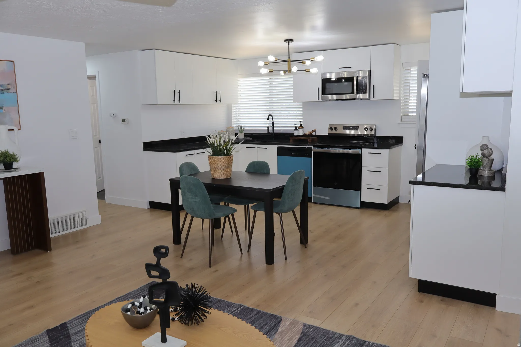 Open-concept kitchen and dining area featuring light wood-finish flooring, white cabinetry with black hardware, black countertops, and stainless steel appliances