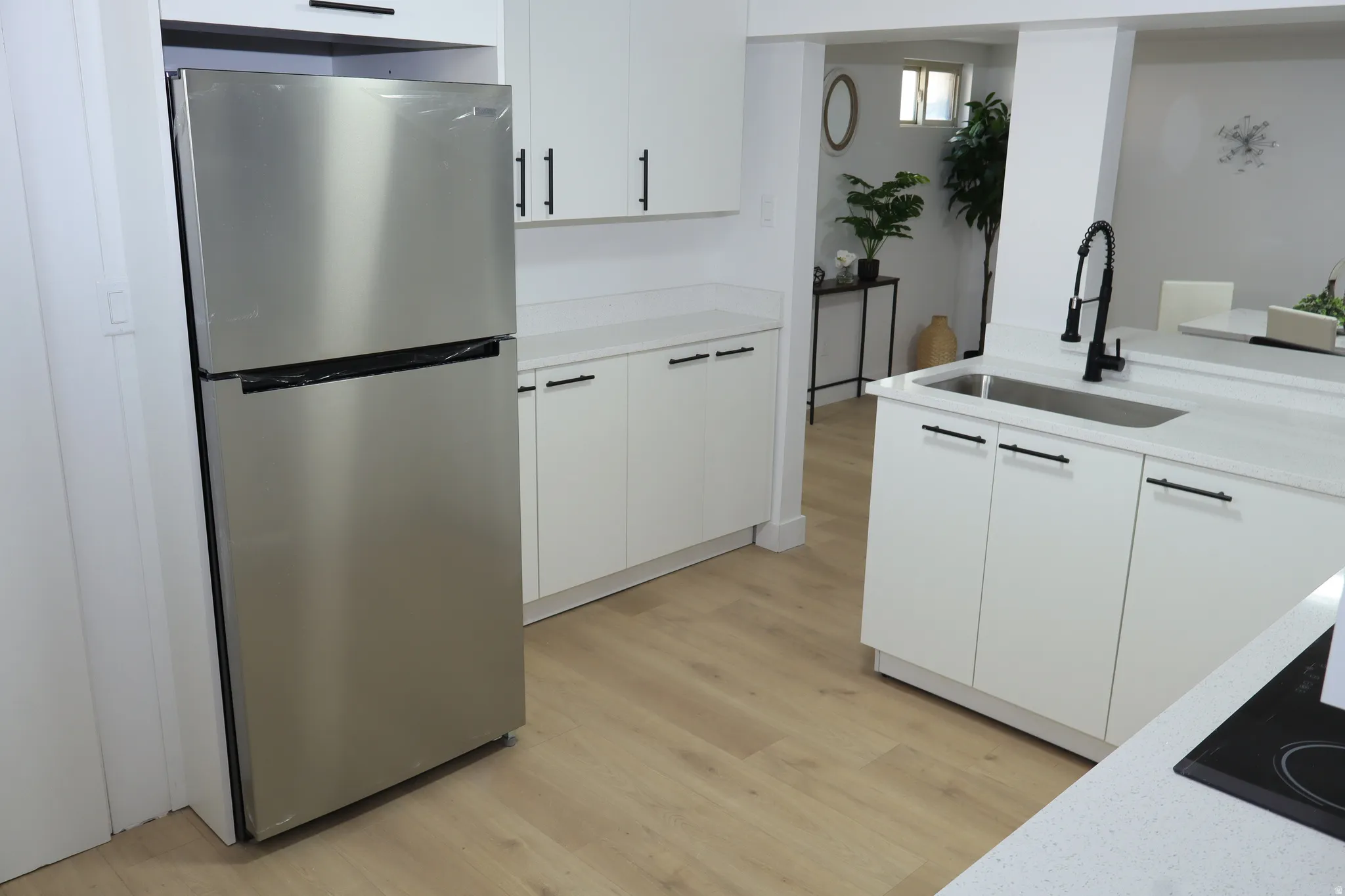 Modern kitchen featuring light wood-finish flooring, white cabinetry with matte black hardware, light-colored countertops, a stainless steel refrigerator, and a kitchen island with an undermount sink and matte black pull-down faucet