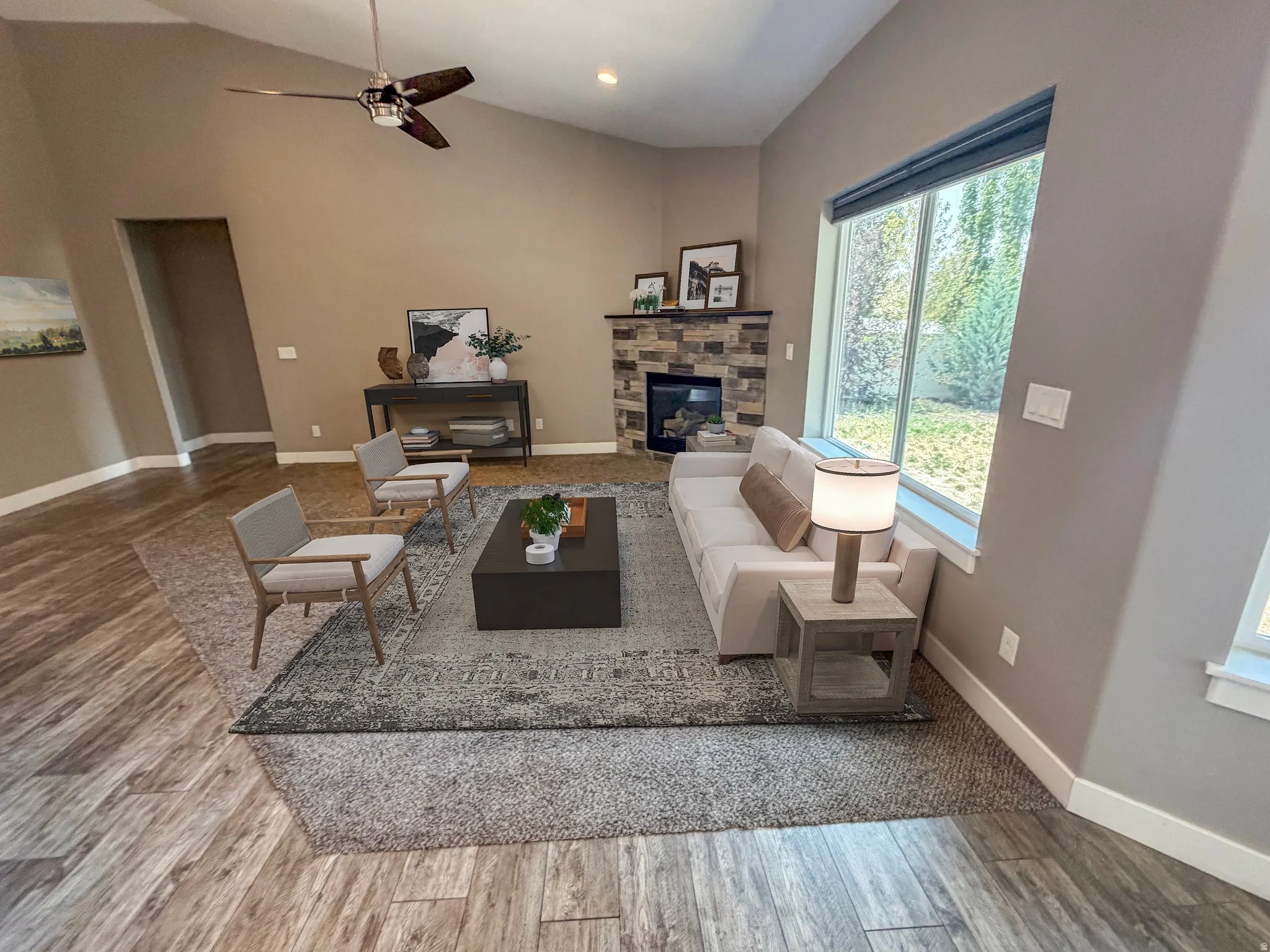 Vaulted ceiling living space featuring a stone fireplace with a built-in firebox, large picture windows, wood-finish flooring, and a ceiling fan with integrated lighting