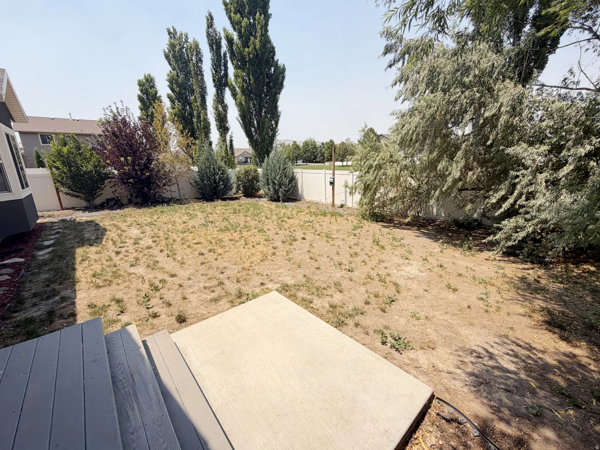 Expansive backyard featuring a gray composite deck, a concrete patio, a full privacy fence, and mature trees and shrubs providing natural screening
