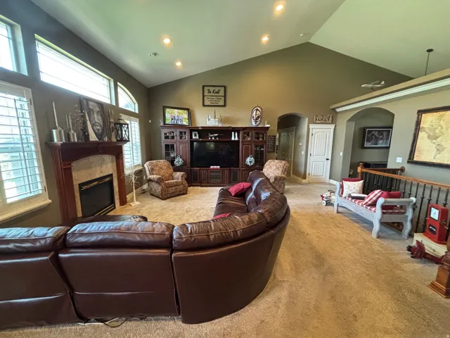 Vaulted ceilings with recessed lighting, large windows with plantation shutters, a fireplace with a stone surround and wood mantel, an arched doorway, and a carpeted floor