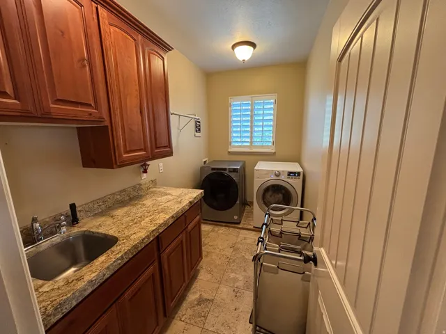 Dedicated laundry room featuring warm wood cabinetry, granite-style countertops, a stainless steel utility sink, tiled flooring, and a window with plantation shutters