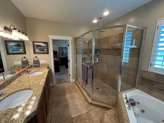 Bathroom featuring a dual vanity with a granite-finish countertop, undermount sinks, a glass-enclosed shower with tiled walls, a separate built-in soaking tub, and tile flooring