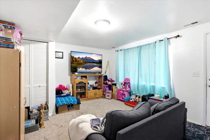Carpeted living area featuring white walls, a built-in closet with louvered bifold doors, a single ceiling light fixture, and a window with a curtain rod installation
