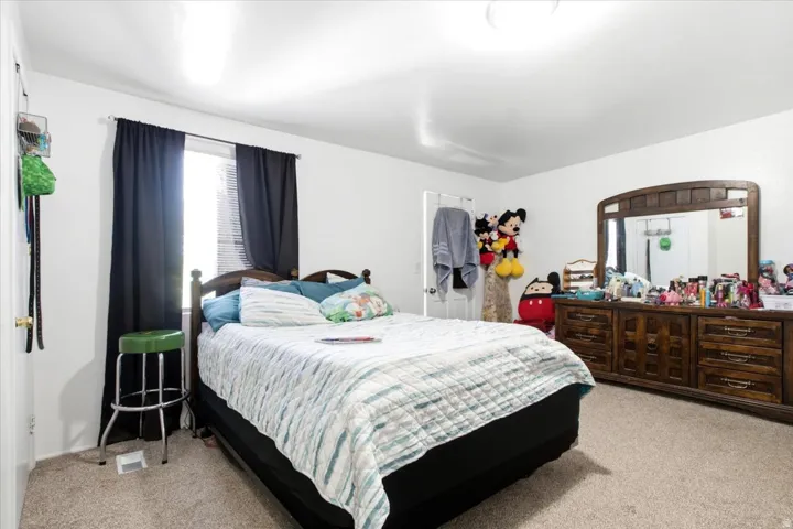 Carpeted bedroom with a window featuring blinds and dark curtains, white walls, and a ceiling light fixture