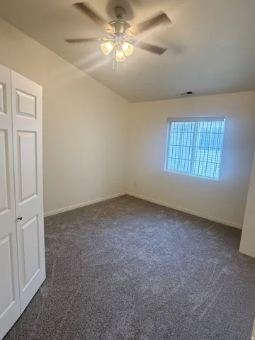Bedroom featuring neutral wall paint, grey patterned carpeting, a multi-blade ceiling fan with integrated lighting, and a window with horizontal blinds and security bars