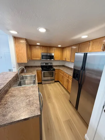 Recessed lighting illuminates the kitchen featuring wood cabinetry, stainless steel appliances, a double basin sink, and wood-finish flooring