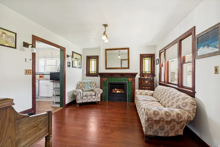 Living area featuring a green tile fireplace with a wood mantel, wood-finish flooring, and natural wood trim around windows and doorways