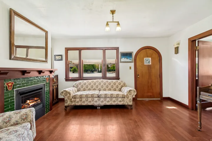Living area featuring hardwood flooring, a fireplace with green tile surround, wood-trimmed windows, a solid wood arched doorway, and a brass ceiling light fixture