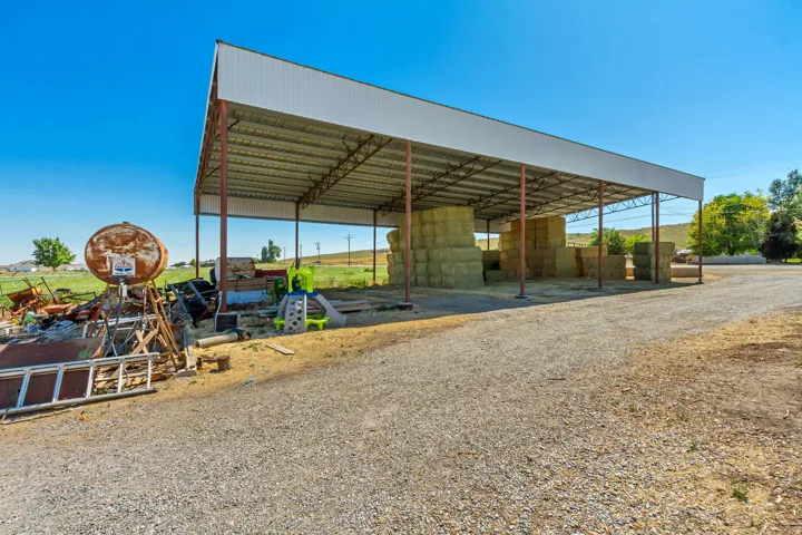 Large open-sided metal structure with a gabled roof and supporting beams, situated on a gravel surface