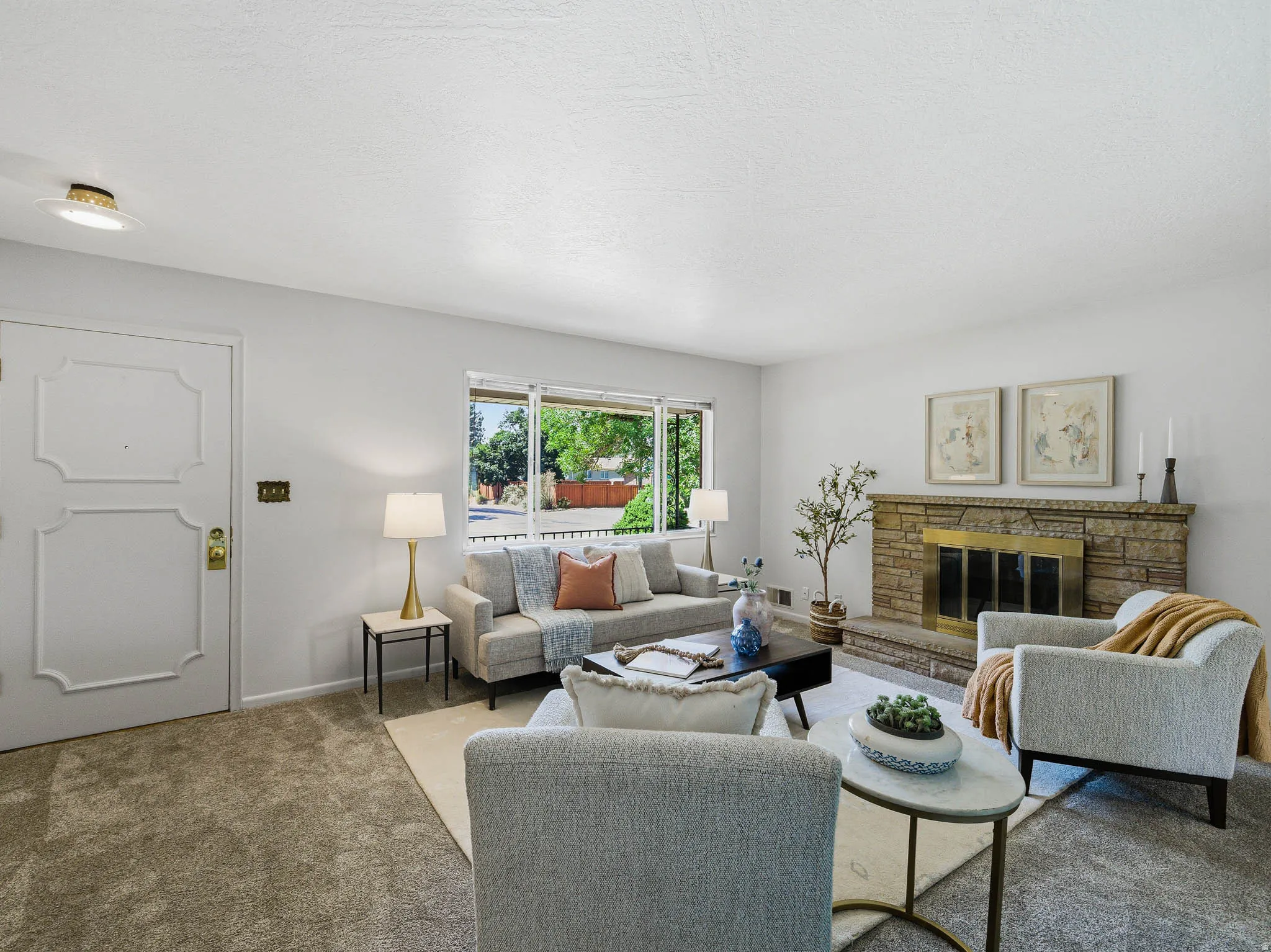 Spacious living room featuring a stone fireplace with brass accents, light carpeting, an expansive window, and a white paneled entry door