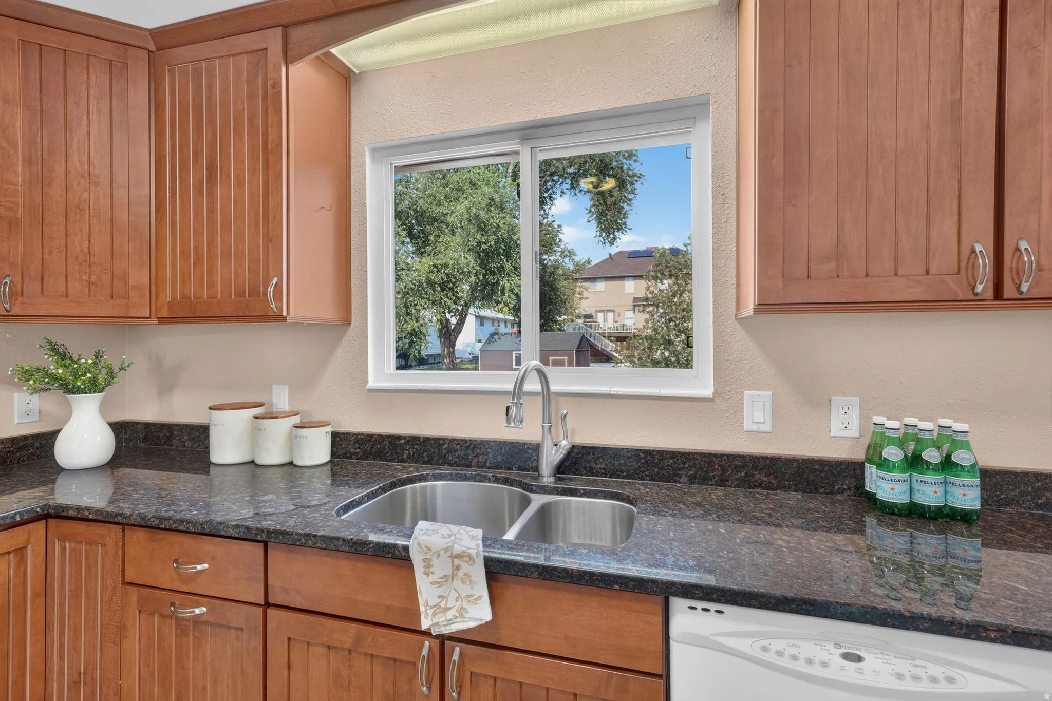 Kitchen featuring a double basin stainless steel sink with a gooseneck faucet, dark speckled countertops, wood cabinetry, and a white dishwasher