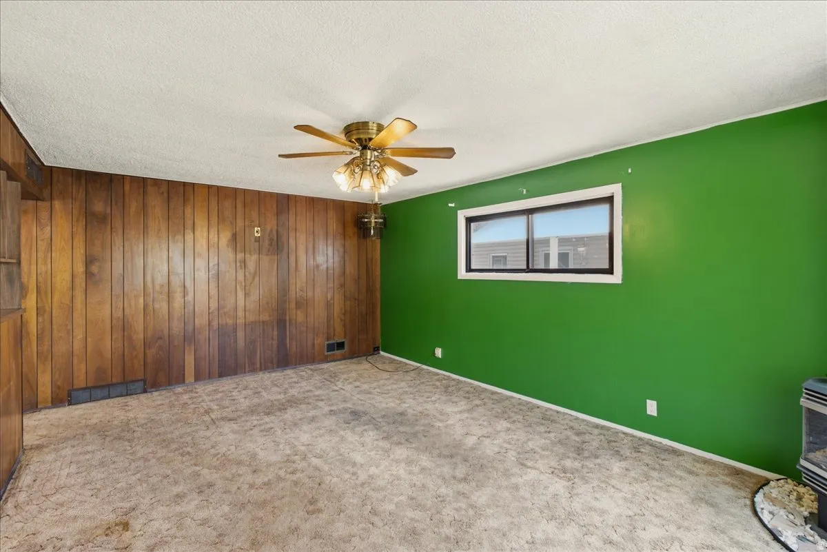 Spacious room featuring wood paneling, a large window, a ceiling fan with integrated lighting, and a floor-mounted heating unit