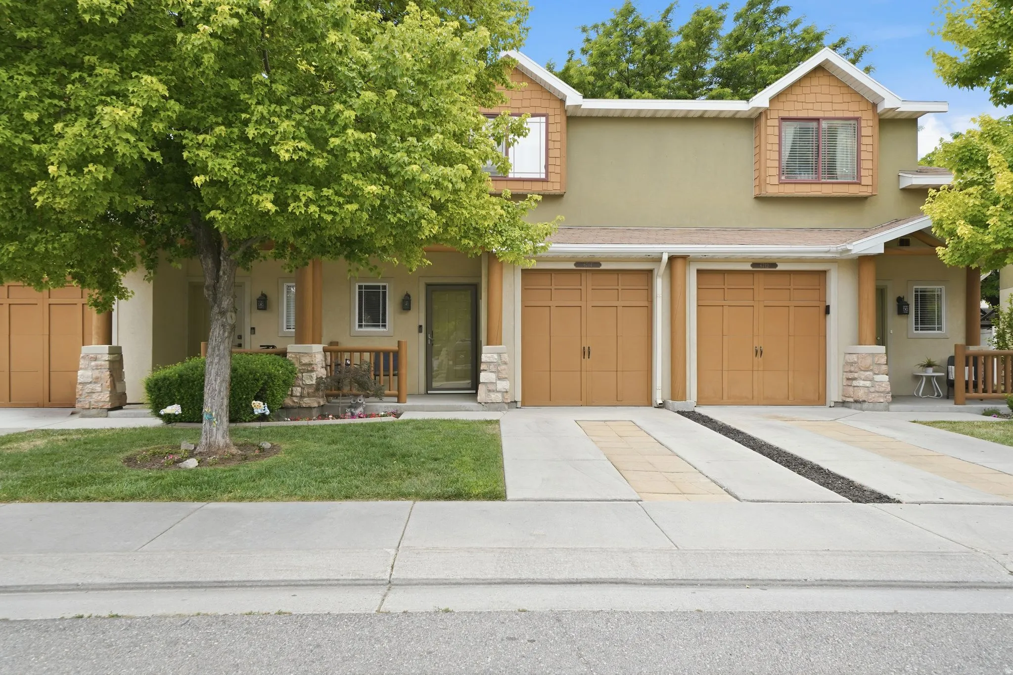 Duplex exterior featuring two garage doors, front entry porch, upper-level dormers with wood siding, stucco facade, and a concrete driveway with decorative stone accents