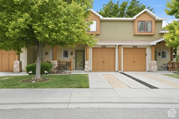 Duplex exterior featuring two garage doors, front entry porch, upper-level dormers with wood siding, stucco facade, and a concrete driveway with decorative stone accents