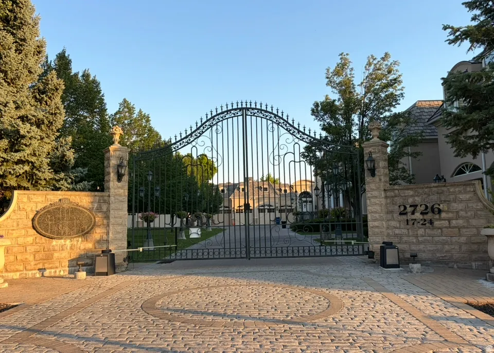 Ornate wrought iron gate set within stone columns, featuring integrated lighting and address numbering, with a brick paver driveway