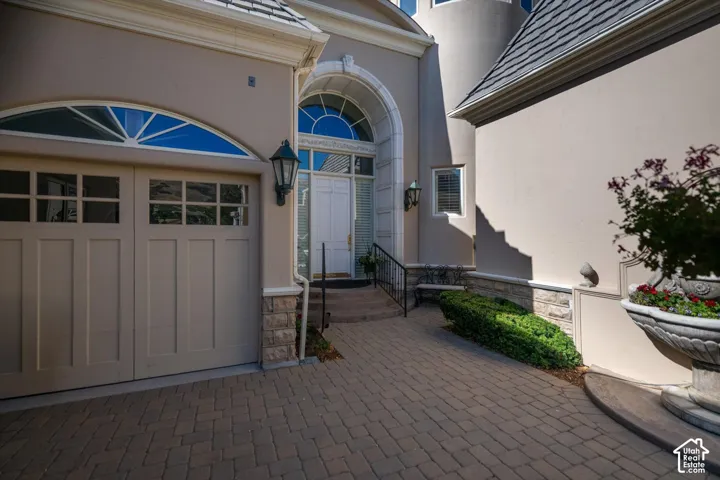 Paver driveway leading to a two-car garage with arched transom window, a grand entry door with sidelights and transom, exterior sconce lighting, and stone veneer accents