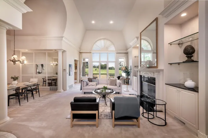 Spacious living area featuring vaulted ceilings, a large arched window, a fireplace with a stone mantel, built-in cabinetry with glass shelving, and light-toned carpeting