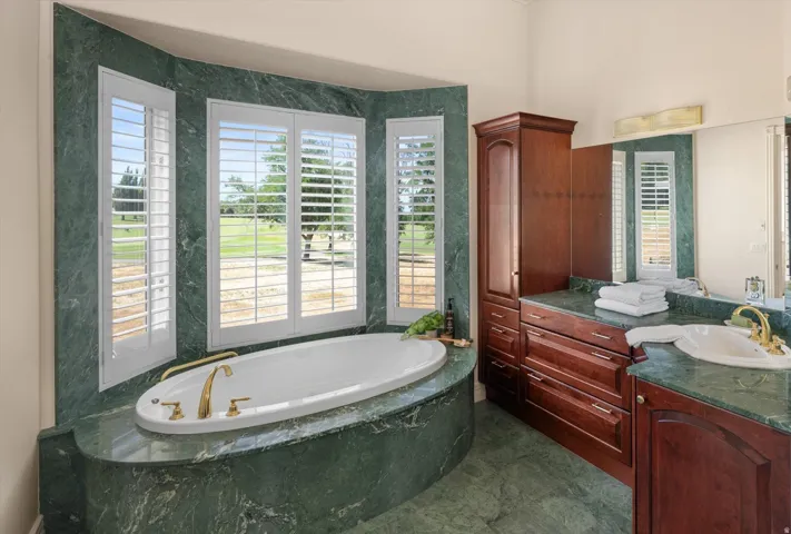 Soaking tub set in a green stone surround with brass fixtures and plantation shutters