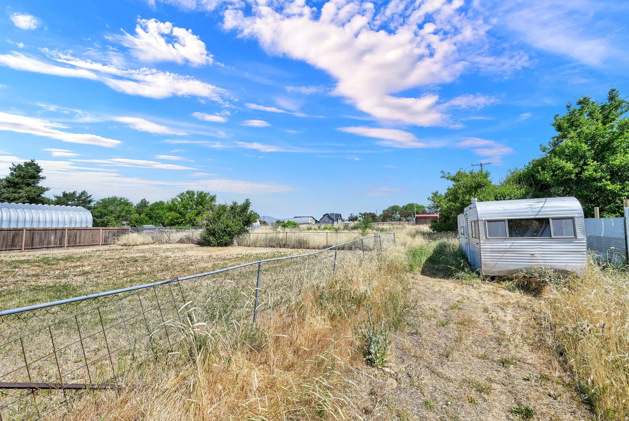 Expansive open lot featuring a chain-link fence, a distant wood fence, and mature trees