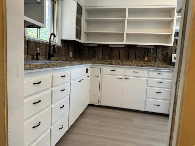 White cabinetry with black hardware, stone-finish countertops, wood-panel backsplash, and light wood-finish flooring
