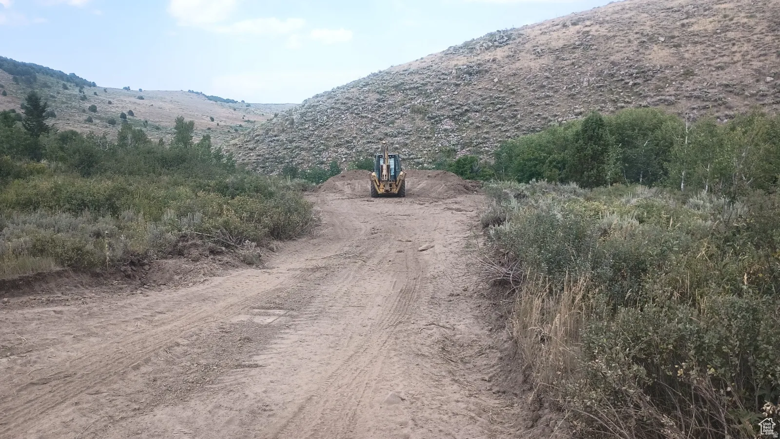 Undeveloped parcel featuring a dirt path, natural scrub brush, and distant tree lines, set against a mountainous backdrop