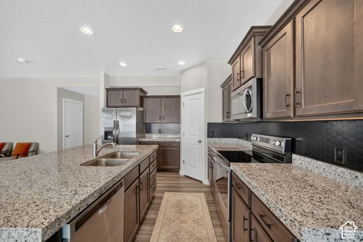 Kitchen featuring granite countertops, wood-finish flooring, dark wood cabinetry, a stainless steel double-basin sink, and a black honeycomb tile backsplash