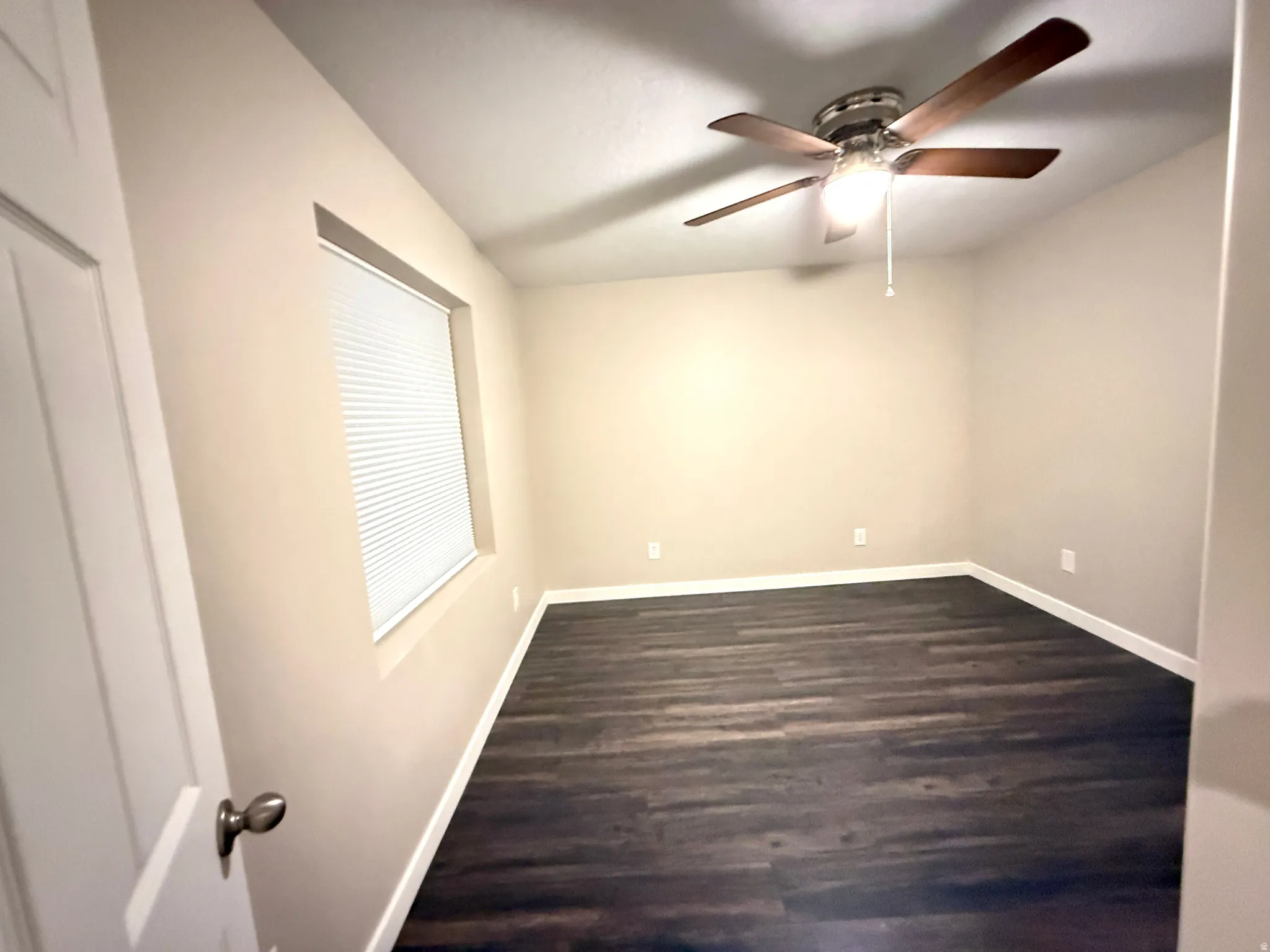 Spacious room featuring wood-finish flooring, a contemporary ceiling fan with integrated lighting, neutral wall tones, and a window with blinds
