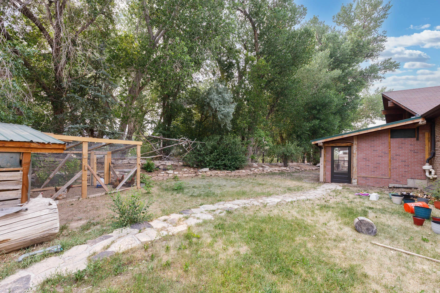 Expansive backyard featuring a stone pathway, mature trees, a wood-framed animal enclosure with a metal roof, and a brick-sided structure with a glass-paneled door