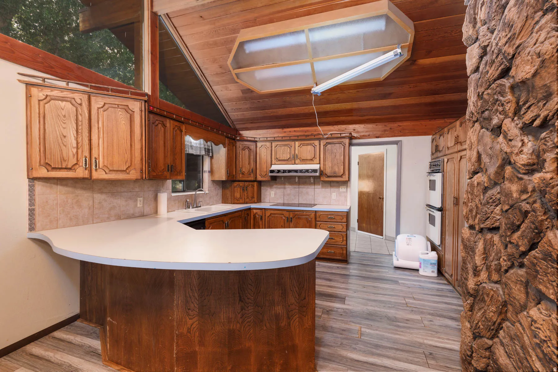Kitchen featuring a wood-panel vaulted ceiling, wood-finish flooring, full suite of built-in appliances, ample wood cabinetry, and an expansive stone-clad fireplace
