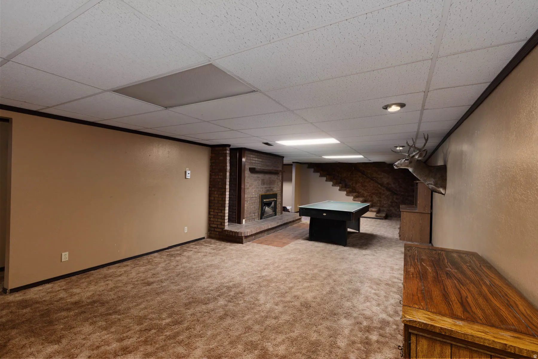 Spacious carpeted room featuring a brick fireplace with a raised hearth, wood-finish staircase, and drop ceiling with recessed lighting
