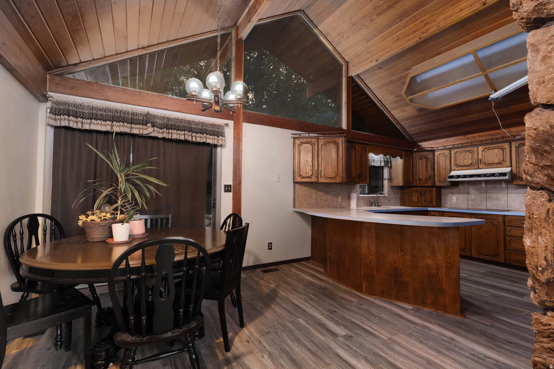 Vaulted wood-paneled ceilings with exposed beams, wood-finish flooring, a kitchen with wood cabinetry and tile backsplash, and a dining area with an overhead chandelier