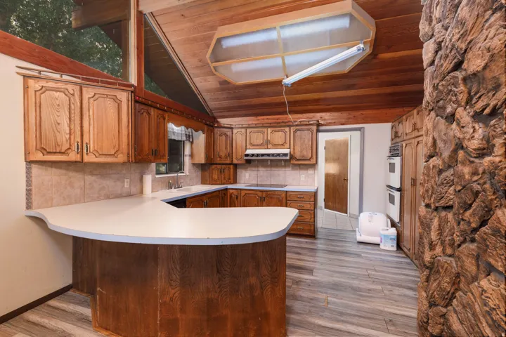 Kitchen featuring a wood-panel vaulted ceiling, wood-finish flooring, full suite of built-in appliances, ample wood cabinetry, and an expansive stone-clad fireplace