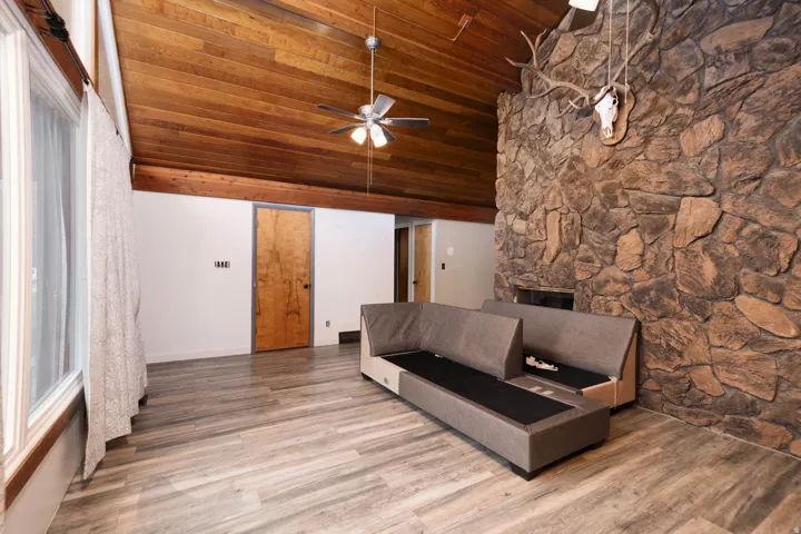 Spacious interior featuring a vaulted wood-paneled ceiling, a substantial stone fireplace, wood-finish flooring, and a ceiling fan with integrated lighting