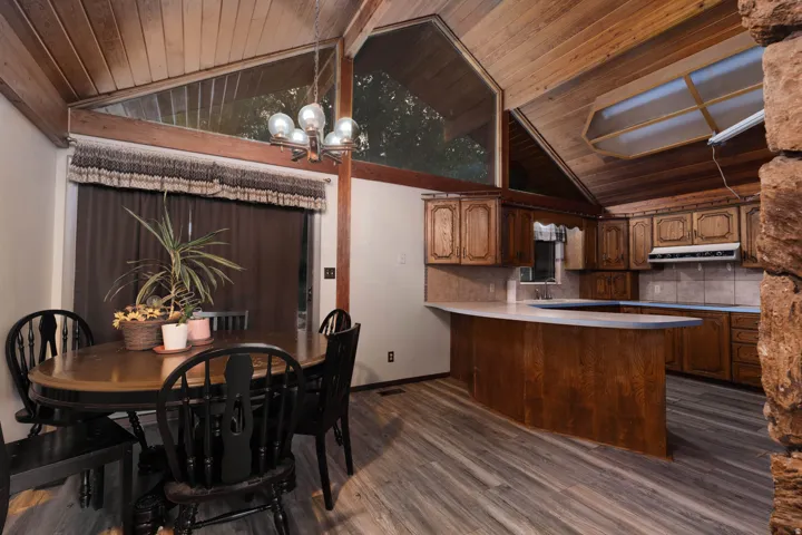 Vaulted wood-paneled ceilings with exposed beams, wood-finish flooring, a kitchen with wood cabinetry and tile backsplash, and a dining area with an overhead chandelier